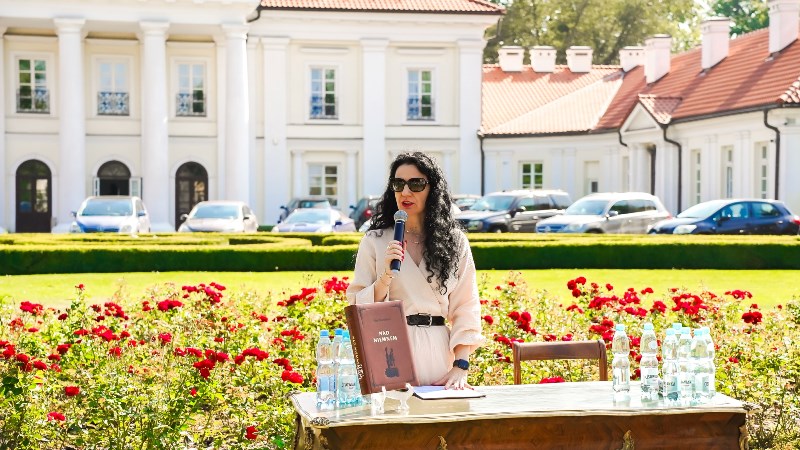 prof. Walęciuk-Dejneka stoi przy stole na którym leży książka Nad Niemnem, przemawia w tle budynku rektoratu